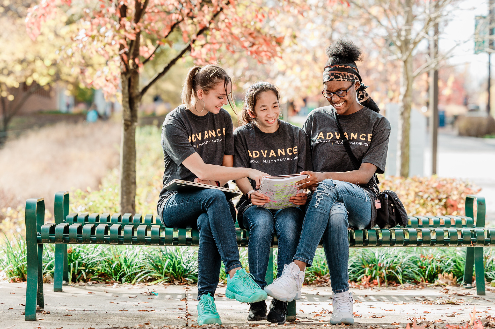 Three college students wearing shirts that say "ADVANCE" smiles as they sit next to each other on a bench.