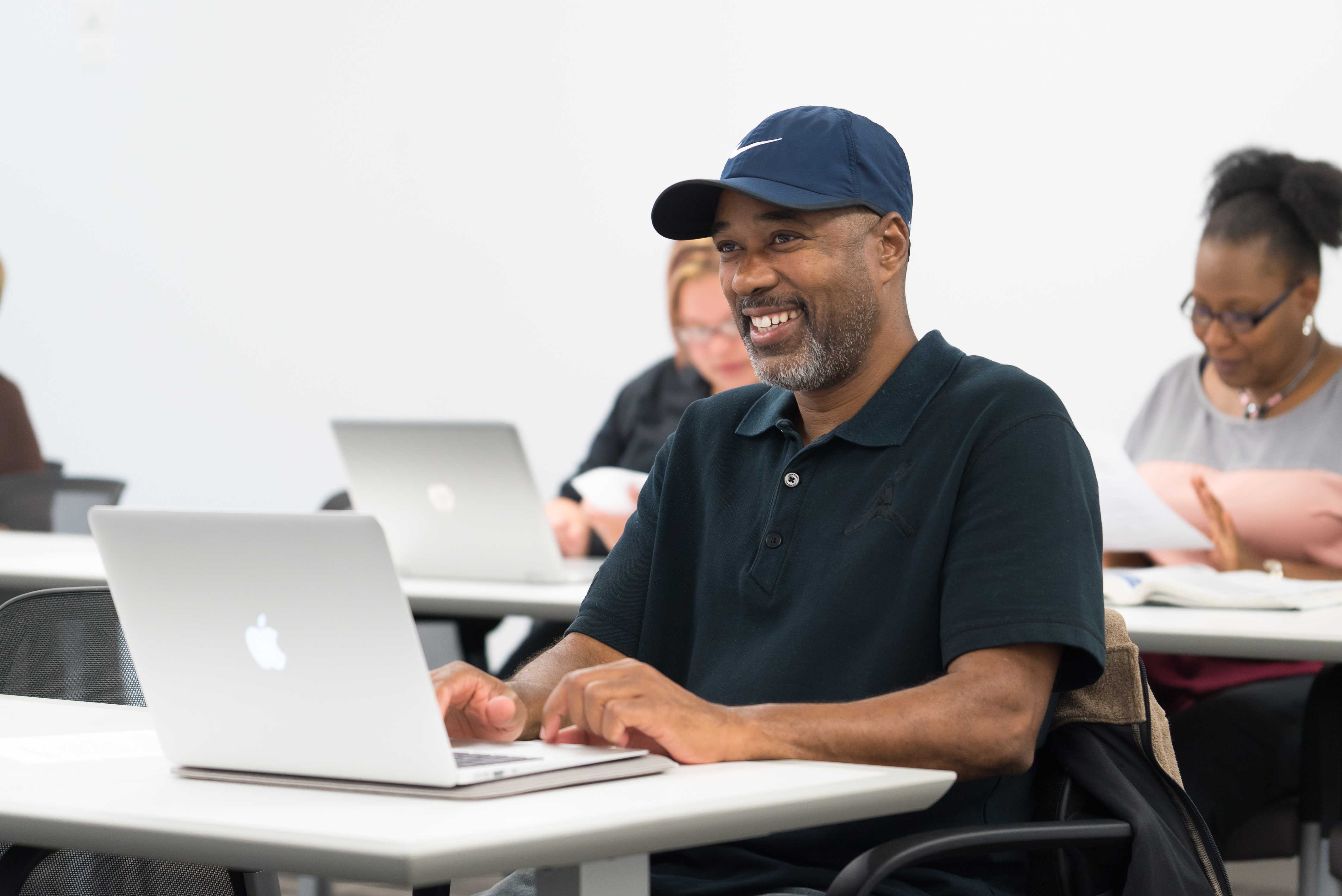 Students sit in a classroom in front of laptops wearing casual clothes, smiling towards an instructor who is off screen. 