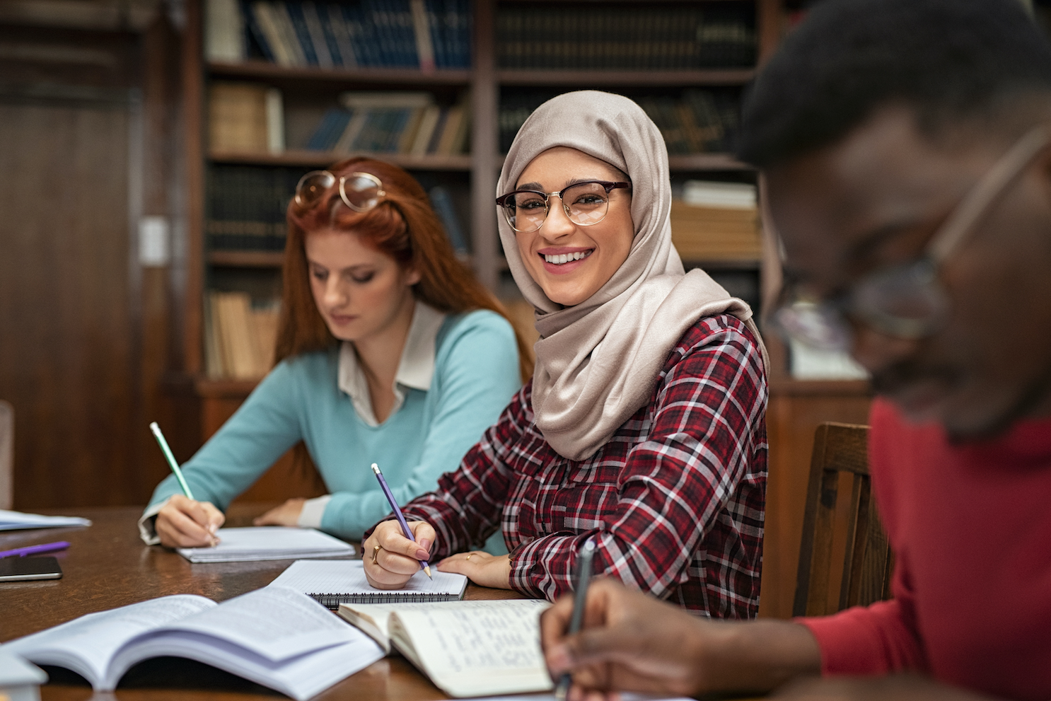 Picture of a young woman in a headscarf taking notes in class and smiling. 