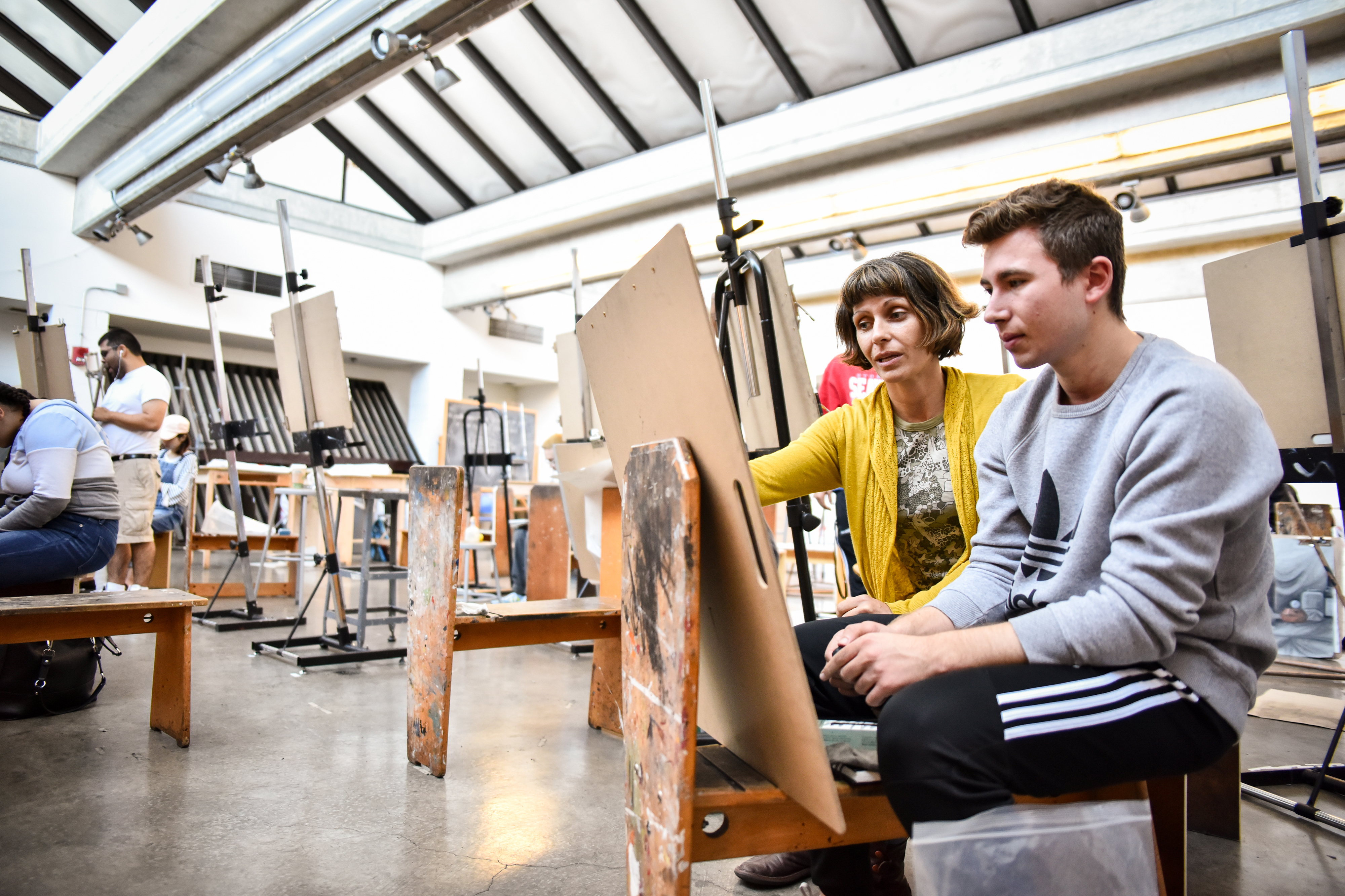 A faculty member oversees a student art project at an easel.
