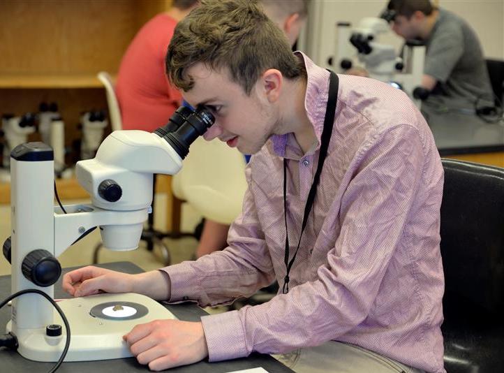 Photo of a teenage boy looking into a microscope in a lab classroom. A few more students are in the background at another table.