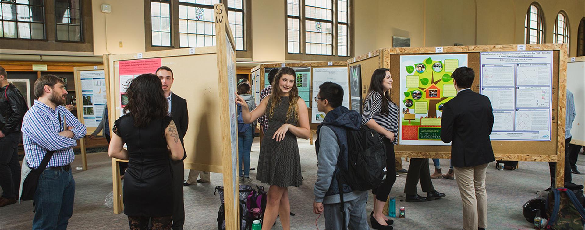 A woman stands in front of academic posters, explaining something to someone standing next to her.