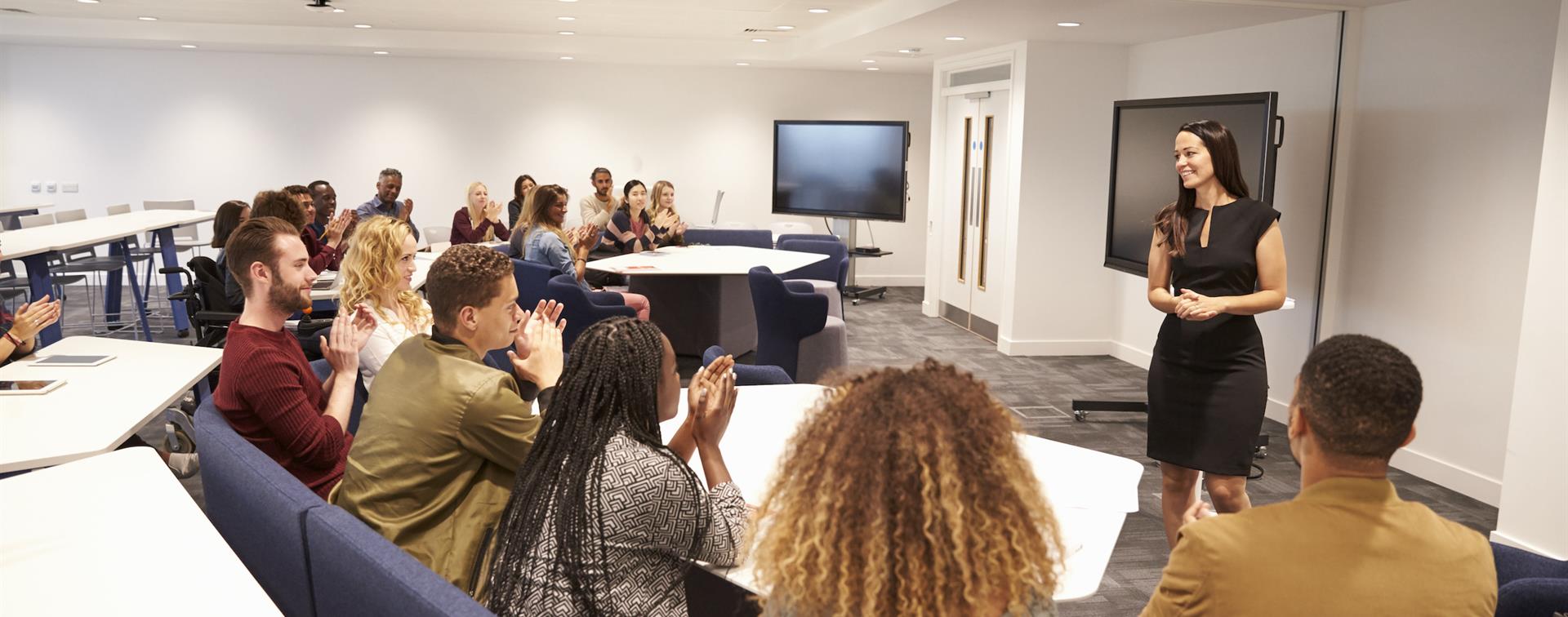 Photo of woman completing presentation in front of classroom as students at desks applaud