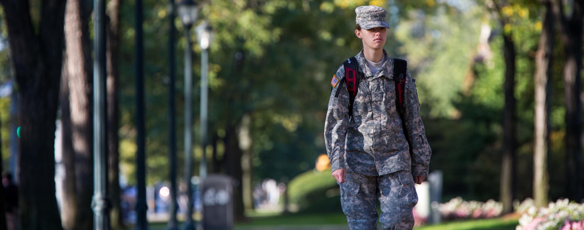A military member walking along a campus sidewalk with a backpack on