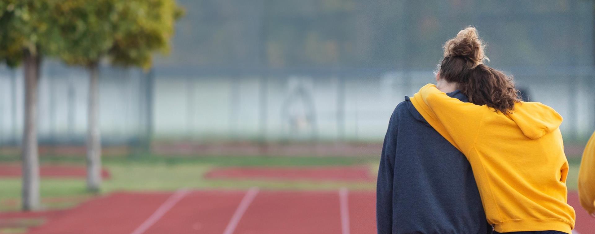 Two individuals stand on a track, holding each other in support while walking away from the camera.