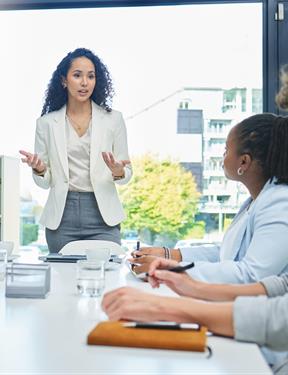 Photo of woman in a suit at the head of a table addressing a group in a modern conference room