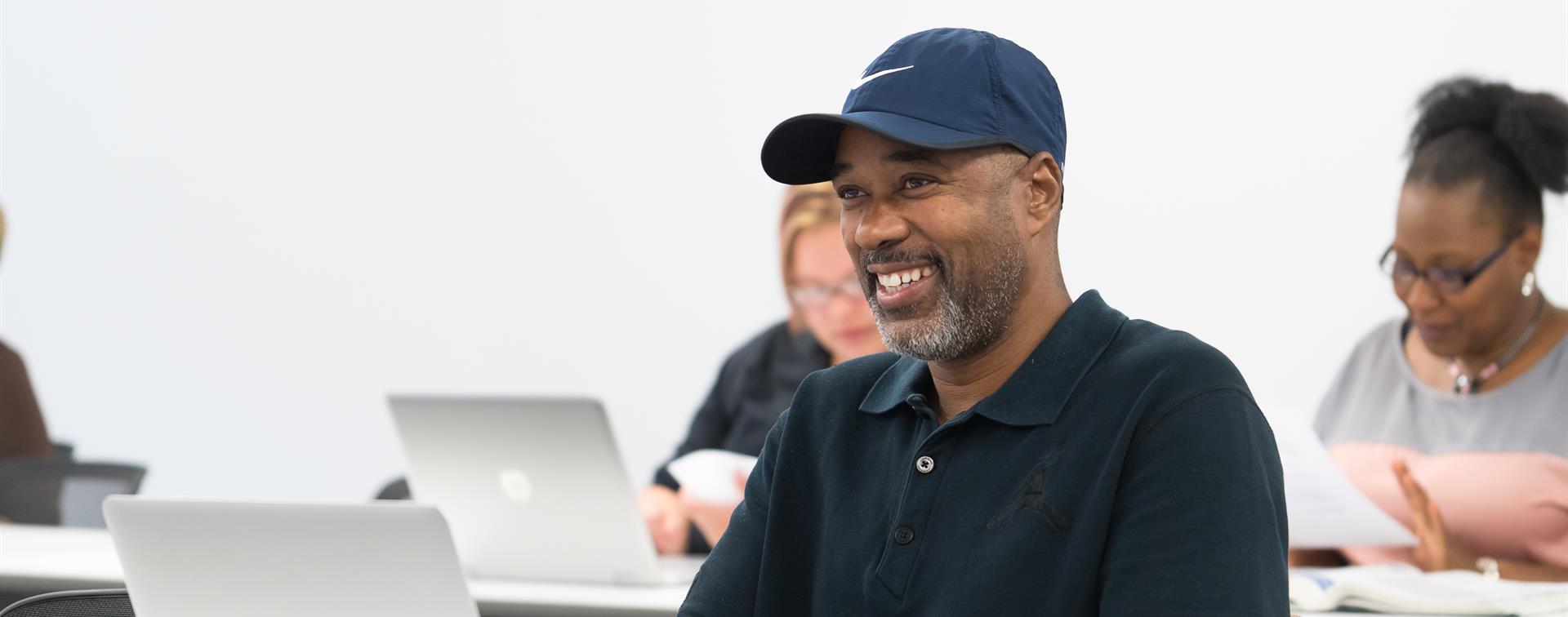 Students sit in a classroom in front of laptops wearing casual clothes, smiling towards an instructor who is off screen. 