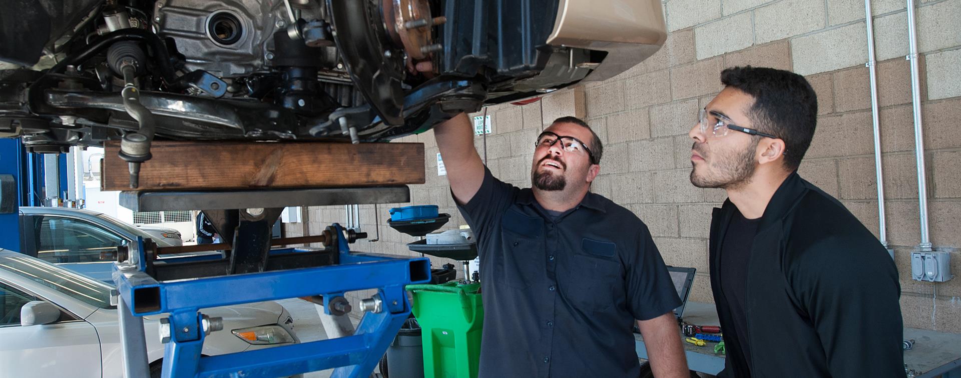 Two individuals stand in a mechanic shop, pointing to the machinery above them on a lift.