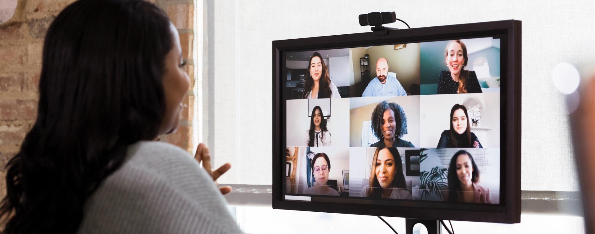 An individual sits at a computer screen wearing headphones while others appear on video during a conference call.