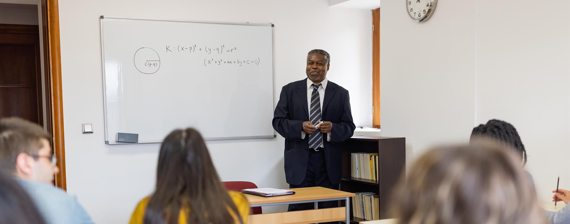 A teacher stands in front of a whiteboard with equations written on it.