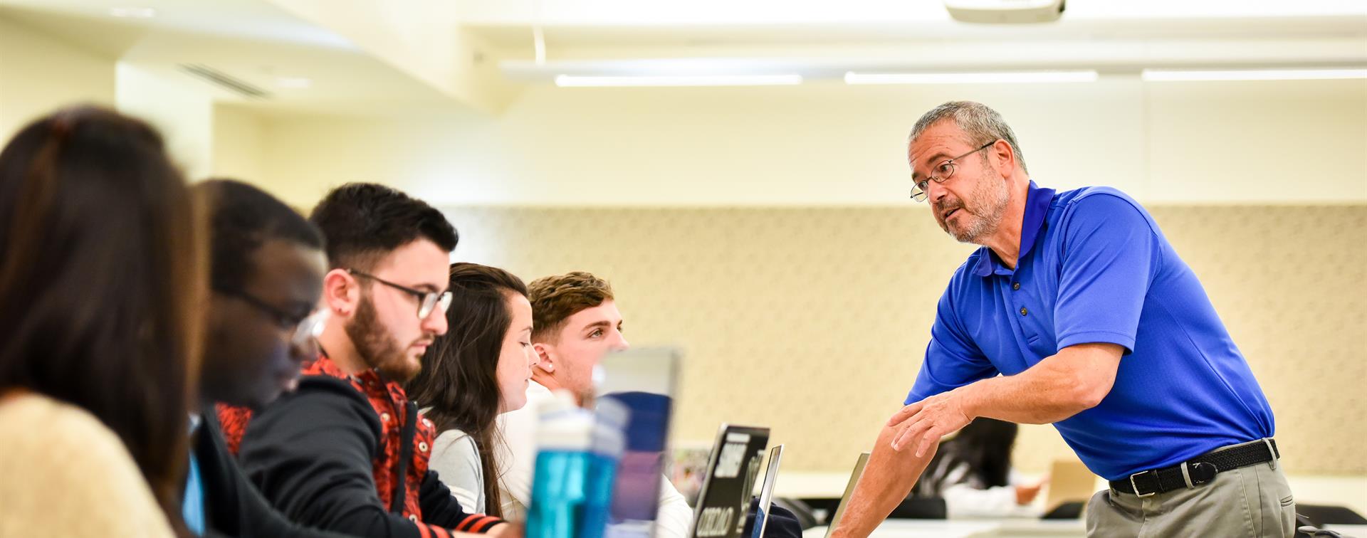 A group of five students in a classroom listen to a lecturer wearing a blue polo shirt