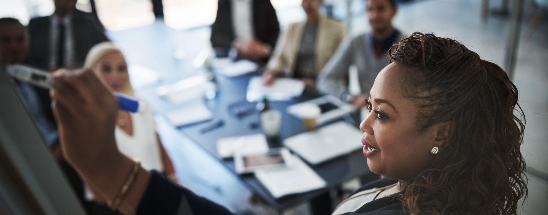 A woman in business attire writes on a whiteboard while presenting to a small group