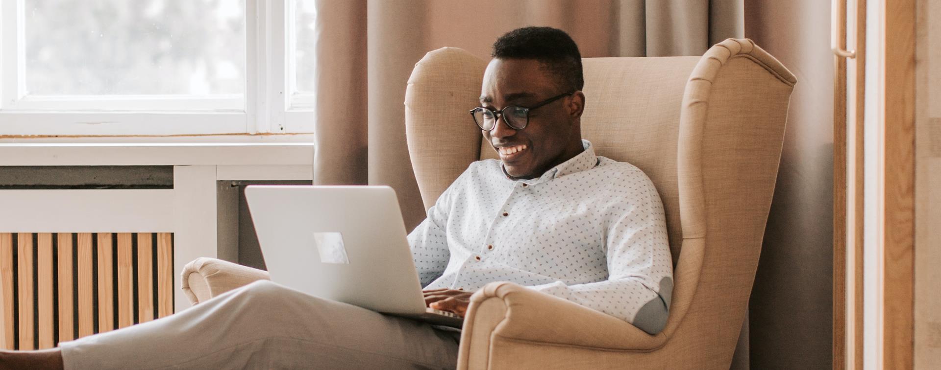 An individual wearing glasses sits in an armchair smiling at a laptop.