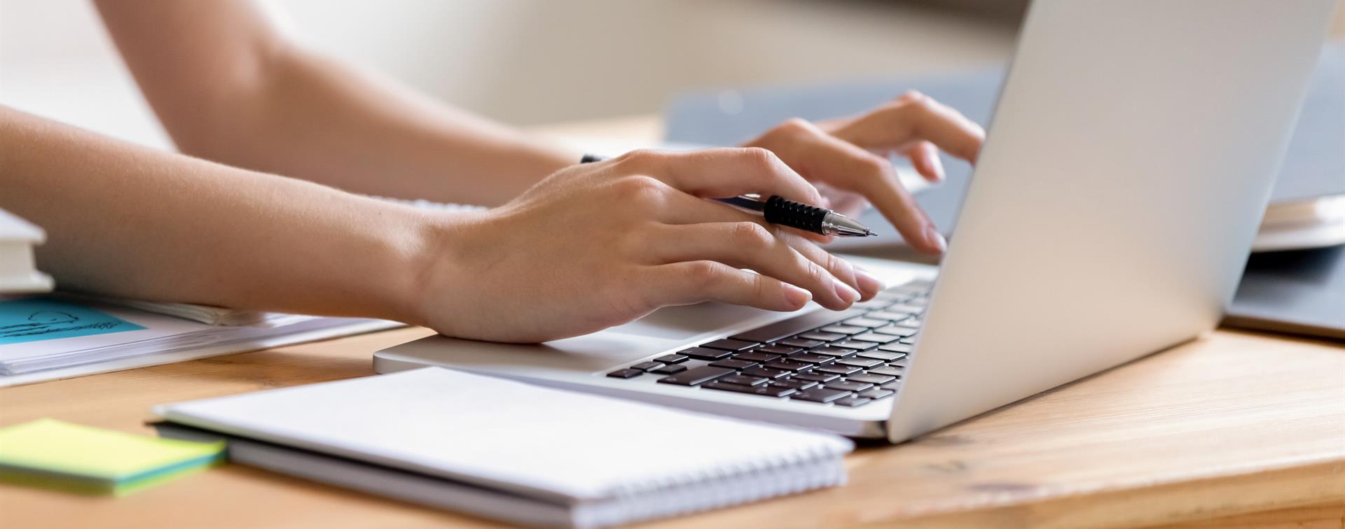 Photo of hands typing on a laptop with open notebook lying in foreground on desk