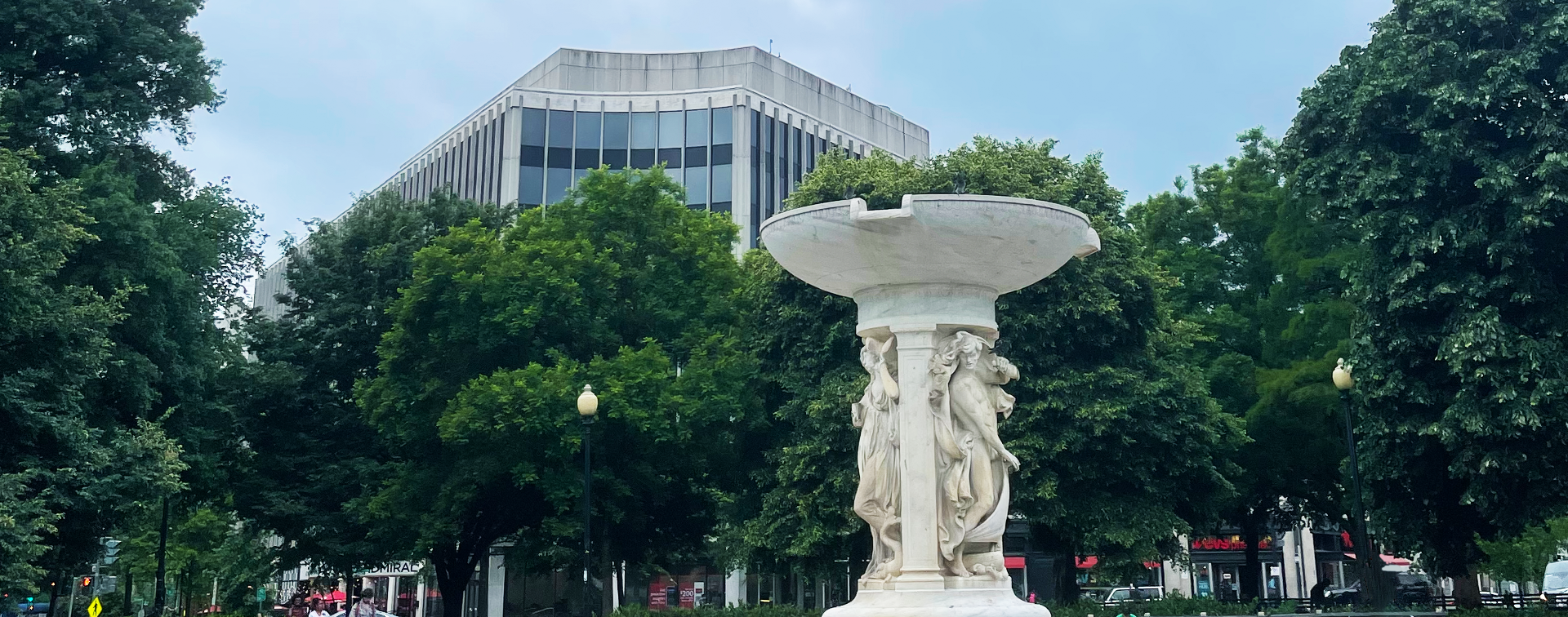 A picture of One Dupont Circle behind a group of green trees and a fountain in the center of Dupont Circle in Washington, DC