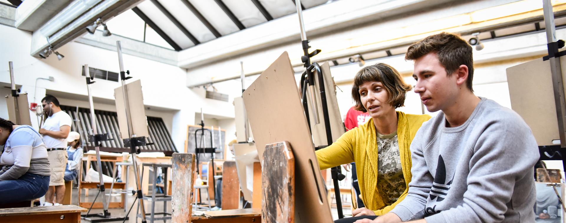 A faculty member oversees a student art project at an easel.