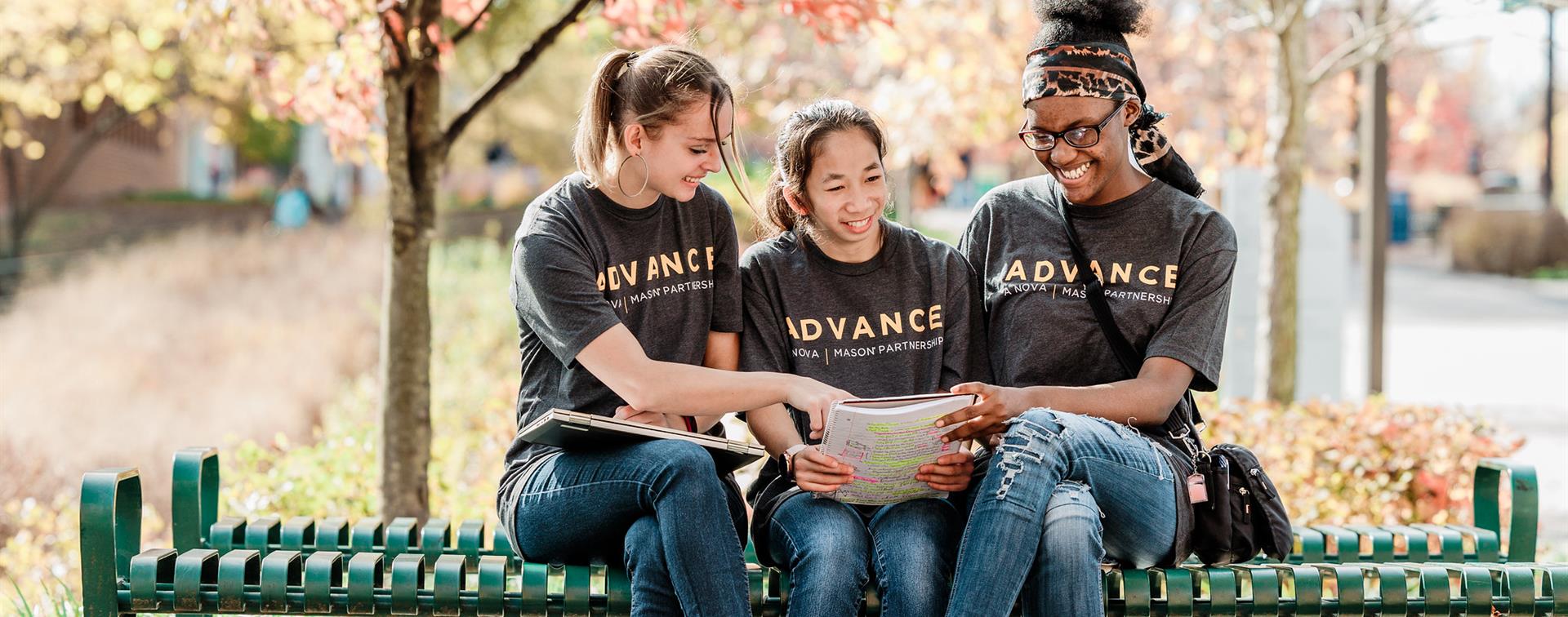 Three college students wearing shirts that say "ADVANCE" smiles as they sit next to each other on a bench.