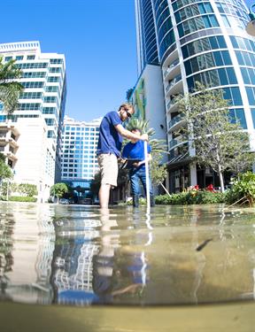 Tall buildings fill the image from Florida Atalantic University. Two people stand in front with water coming up to their ankles.