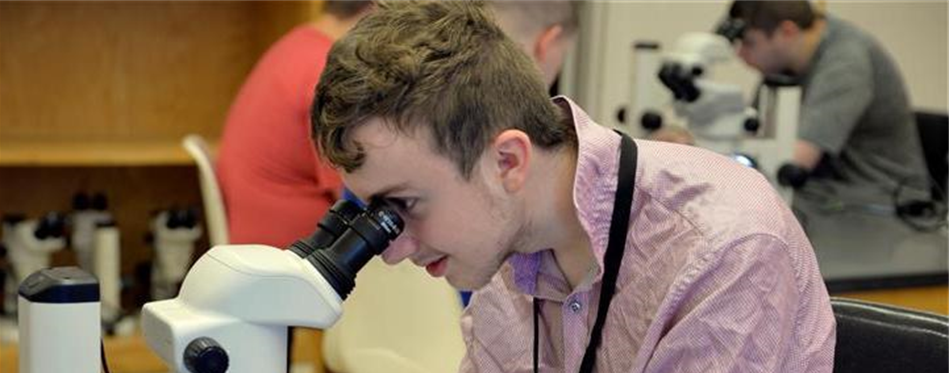 Photo of a teenage boy looking into a microscope in a lab classroom. A few more students are in the background at another table.