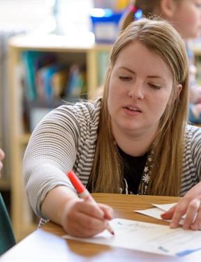 An educator sits as a desk with a pen in hand, activley intructing someone sitting next to them off screen.