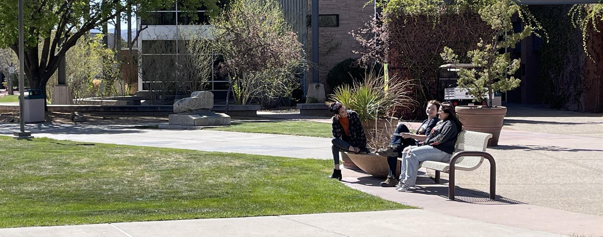 Photo of three college students sitting and chatting on a bench on a campus. Plants and trees bloom behind them.
