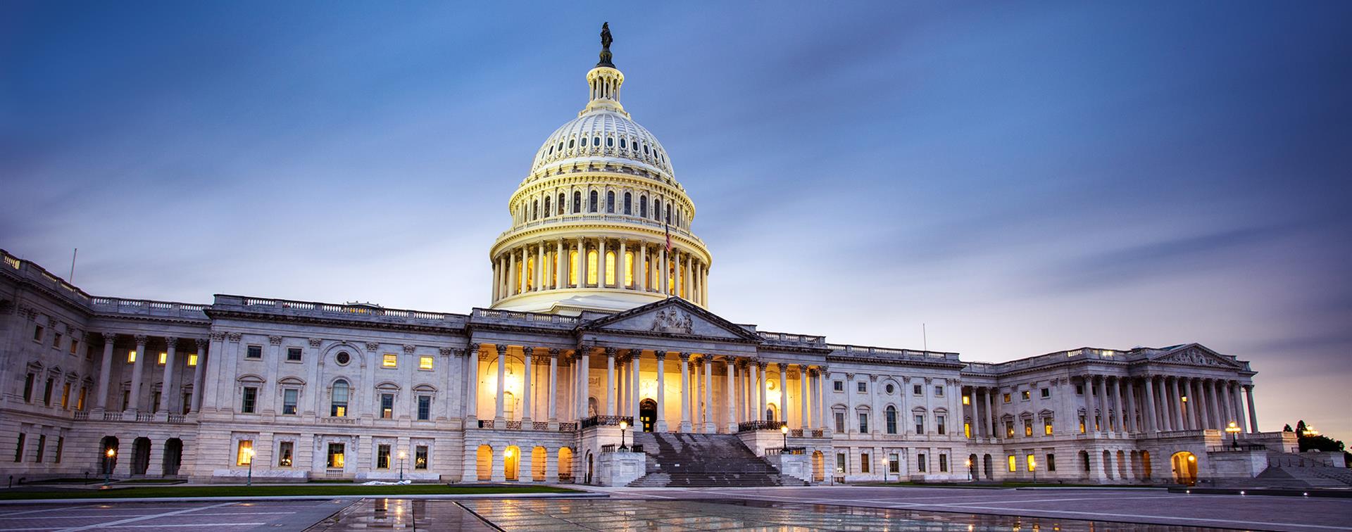 A picture of the US Capitol building, all the windows are illuminated and the ground is wet from rain. It is either dusk or dawn