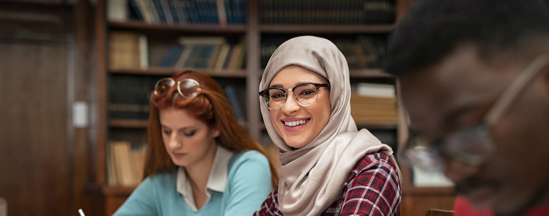 Picture of a young woman in a headscarf taking notes in class and smiling. 